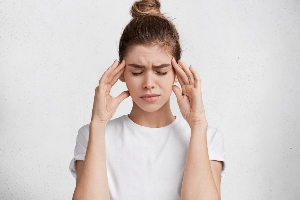 Młoda kobieta w białej koszulce trzyma się za skronie z wyrazem bólu twarzy. Zdjęcie ilustruje ból głowy lub migrenę.